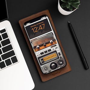 Può includere: Una custodia per telefono in pelle marrone con un design radio vintage. Lo schermo del telefono mostra un orologio digitale con l'ora 12:47 e la data sabato 10 febbraio.