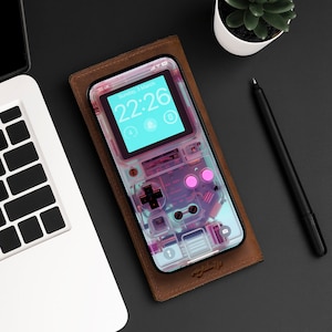 May include: A brown leather phone case with a clear, transparent phone inside. The phone screen displays a digital clock with the time 22:26 and the date Sunday, 3 March. The phone case has a pink button and a clear, transparent design that resembles a retro video game console.