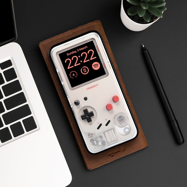 May include: A brown leather case with a white Game Boy Color style phone case. The phone case has a digital clock displaying "Sunday, 3 March 22:22" and the word "GAMEBOY" printed on it.
