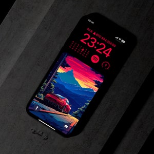 Op de afbeelding: Een rode sportwagen rijdt over een kronkelende weg met een bergketen op de achtergrond. De auto rijdt richting een zonsondergang met een roze, oranje en blauwe lucht. De afbeelding wordt weergegeven op het scherm van een smartphone met een digitale klok die 23:24 uur aangeeft en een Bitcoin-prijs van € 44.224,99.