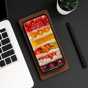 Op de afbeelding: Een bruine leren telefoonhoes met een helder telefoonscherm waarop een gelaagde cake met fruit en slagroom te zien is. Het telefoonscherm toont de tijd 16:52 en de datum zo 28.