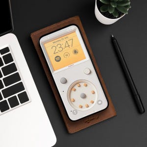 May include: A brown leather case with a white phone inside. The phone has a digital clock displaying "23:47" and the text "Monday 22 July". The phone also has a circular control panel with multiple buttons.