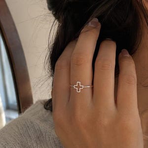 May include: A silver ring featuring a minimalist cross design. The ring is thin and delicate, with the cross shape open in the center. The ring is worn on a finger, and the overall aesthetic is simple and elegant.