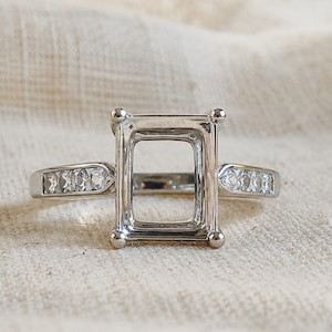 May include: A silver-toned metal ring setting featuring a rectangular center mount with four prongs designed to hold a gemstone. The band is accented with small, clear, round stones embedded along both sides of the shank, creating a polished look.