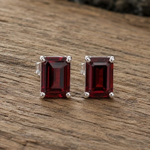 May include: A pair of stud earrings featuring rectangular emerald-cut deep red garnet gemstones set in polished silver-toned metal. Each gemstone is secured by four small prongs at the corners, showcasing a faceted surface that reflects light.