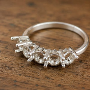 May include: A silver ring setting featuring five empty prong settings arranged in a row along the top of the band. The metal has a smooth, polished finish, and the design is intended to hold five individual stones in a secure, elevated position.