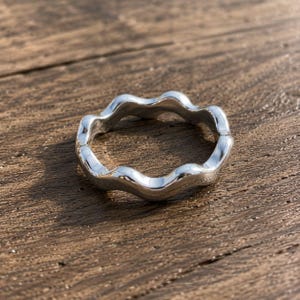 May include: A polished silver-toned ring featuring a continuous wavy, undulating band design. The metal surface is smooth and reflective, creating a series of rounded peaks and valleys that form a circular shape with a consistent thickness throughout.