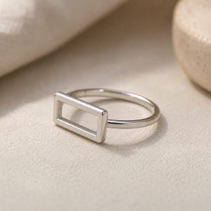 May include: A silver ring with a rectangular open design. The ring band is thin and smooth, leading to the geometric shape. The ring is set against a neutral background, highlighting its modern and minimalist style.