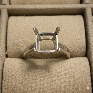 May include: A silver-toned metal ring setting featuring a square-shaped head with four prongs designed to hold a gemstone. The band is smooth and polished, showcasing a classic solitaire style mount ready for a stone to be set into the center.