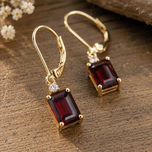 May include: A pair of gold leverback earrings featuring rectangular emerald-cut deep red garnet gemstones. Each earring is accented with a single small round clear white stone set above the garnet, held in a four-prong gold setting.