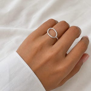 Op de afbeelding: Een gepolijste zilveren ring met een glad, open ovaal ontwerp. De band is dun en cirkelvormig en verbindt zich met een groter, langwerpig ovaal element dat horizontaal over de vinger ligt. Het metaal heeft een heldere, reflecterende afwerking.