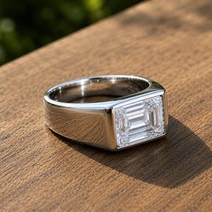 May include: A polished silver-toned metal ring featuring a large, rectangular emerald-cut clear gemstone set horizontally in a wide, smooth band. The ring has a clean, reflective surface and a prominent, faceted center stone with sharp edges.