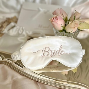 Peut inclure: Masque de sommeil en satin blanc avec le mot "Bride" brodé en or rose.