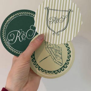 Può includere: Tre sottobicchieri decorativi con design eleganti. Uno è verde scuro con una scritta bianca "R&S", un altro è a righe con uno stemma e "R&S", e il terzo ha un'illustrazione di ombrello e il testo "The Howarths".