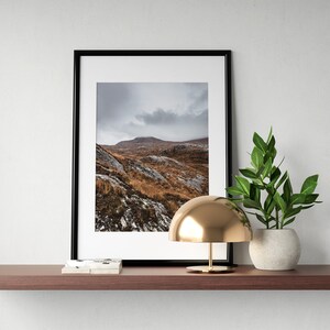 K&ouml;nnte beinhalten: Gerahmter Landschaftsdruck mit einer Berglandschaft mit braunem und grauem Gel&auml;nde unter einem bew&ouml;lkten Himmel. Der Druck befindet sich in einem schwarzen Rahmen und liegt auf einem braunen Regal mit einer goldenen Lampe und einer Topfpflanze.
