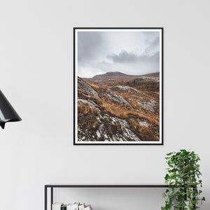 K&ouml;nnte beinhalten: Ein gerahmter Landschaftsdruck mit einer Bergkette unter einem bew&ouml;lkten Himmel. Der Vordergrund zeigt felsiges Gel&auml;nde mit braunem Gras und Schneeflecken. Das Kunstwerk wird an einer wei&szlig;en Wand ausgestellt.