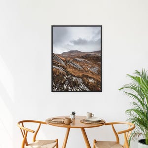 K&ouml;nnte beinhalten: Ein gerahmter Landschaftsdruck mit einer Bergkette unter einem bew&ouml;lkten Himmel. Das Gel&auml;nde weist braune, orange und graue Farbt&ouml;ne mit Schneeflecken auf. Das Kunstwerk h&auml;ngt &uuml;ber einem Holztisch mit zwei St&uuml;hlen.