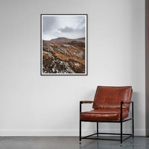 K&ouml;nnte beinhalten: Gerahmte Landschaftsfotografie, die eine bergige Landschaft unter einem bew&ouml;lkten Himmel zeigt. Der Vordergrund zeigt felsige H&uuml;gel mit Flecken aus braunem Gras und Schnee. Rechts befindet sich ein brauner Ledersessel mit schwarzem Rahmen.