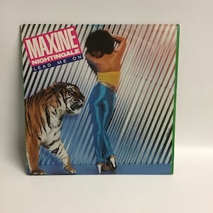 May include: A vinyl record cover featuring a woman in blue pants and a yellow top, with a tiger beside her. The cover has the text "Maxine Nightingale Lead Me On" in pink and white.