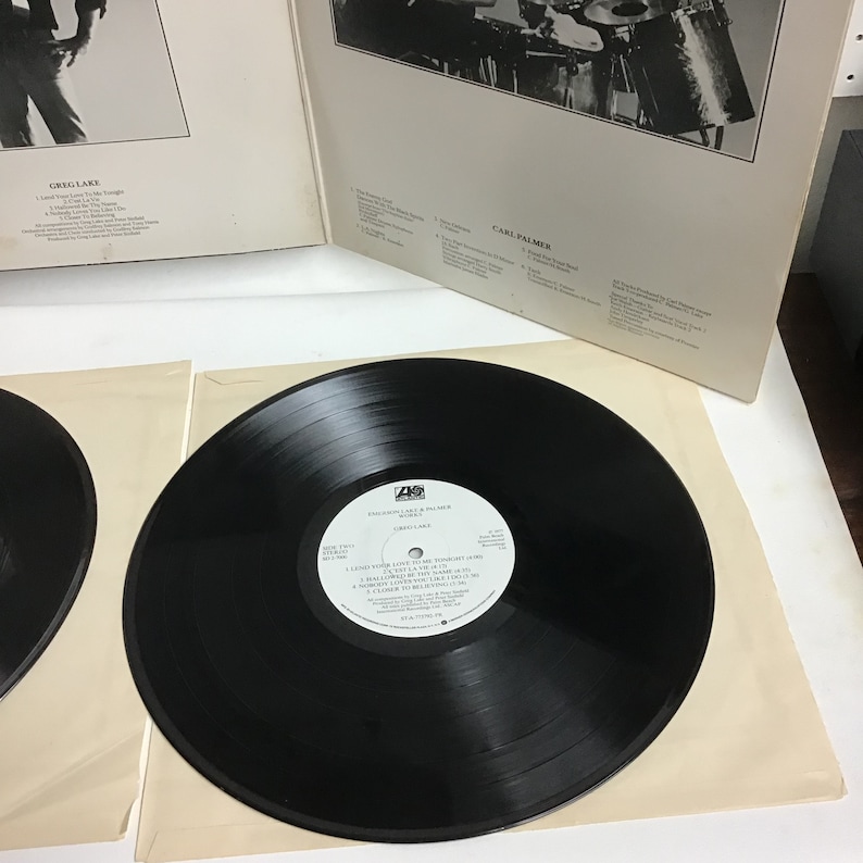 K&ouml;nnte beinhalten: Eine schwarze Schallplatte mit einem wei&szlig;en Label. Das Label tr&auml;gt den Text "Emerson, Lake & Palmer" und "Greg Lake". Die Schallplatte liegt auf einer wei&szlig;en Papieroberfl&auml;che.