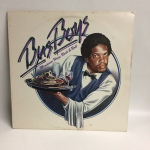 May include: Album cover for the Bus Boys, featuring a man in a waiter's uniform holding a tray with food and drinks. The album title is in a stylized font with the text "Minimum Wage Rock & Roll". The background is a light cream color.