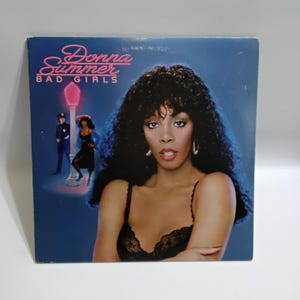 May include: Album cover for Donna Summer's "Bad Girls." The album art features a portrait of Donna Summer with dark curly hair and a black lace top. The album title and artist name are in pink and white text.