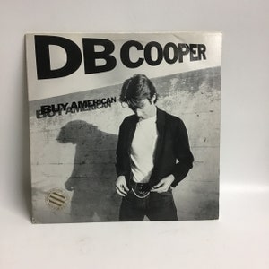 May include: Black and white album cover featuring a man in a black jacket and white t-shirt. The album title is "DB COOPER BUY AMERICAN" with a promotional copy sticker.