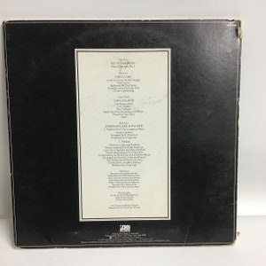 K&ouml;nnte beinhalten: Die R&uuml;ckseite eines Vinyl-Schallplattenalbums f&uuml;r Emerson, Lake & Palmers Album "Fanfare for the Common Man". Das Albumcover ist schwarz mit wei&szlig;em Text, der die Songs auf jeder Seite der Schallplatte auflistet. Der Text enth&auml;lt die Namen der Bandmitglieder Keith Emerson, Greg Lake und Carl Palmer sowie die Songtitel. Das Albumcover enth&auml;lt auch Credits f&uuml;r die Musiker, Produzenten und Toningenieure.