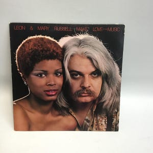 May include: A vintage vinyl record album cover featuring a black woman with short curly hair and a white man with long gray hair. The album title is "Leon & Mary Russell Make Love to the Music".