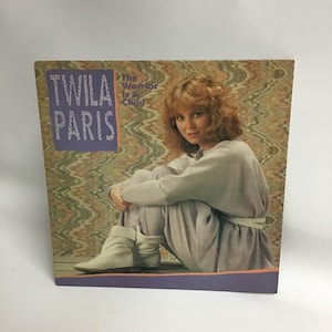May include: A vinyl record album cover featuring a photo of Twila Paris, a female singer-songwriter. The album title is "The Warrior Is A Child" and is printed in purple and white text. The background is a swirling pattern of green, brown, and white.