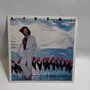 May include: Album cover featuring the artist Airto in a light blue suit, standing barefoot. The background shows a blue sky and water with red flowers. Text on the cover reads "AIRTO" and "I'M FINE / HOW ARE YOU?"