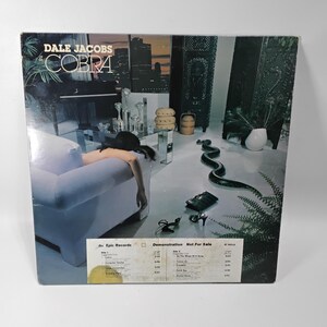 May include: A vintage vinyl record album cover for "Cobra" by Dale Jacobs. The cover features a surreal image with a person reclining on a white sofa, a snake, and city skyline. The album is from Epic Records.