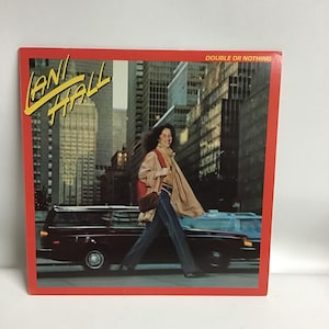 May include: A vinyl record cover featuring a woman walking down a city street. The cover is red with the text "Lani Hall" and "Double or Nothing" in yellow. The woman is wearing blue jeans, a red sweater, and a tan scarf. She is carrying a brown purse.