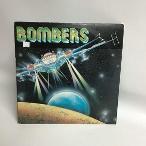 May include: A vintage vinyl record album cover featuring a spaceship flying over a planet with the word "BOMBERS" in large, bold letters.