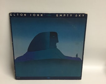 May include: A vintage vinyl record album cover featuring a blue and green illustration of a large, stylized head with a small figure standing on its hand. The album title "Elton John Empty Sky" is printed in white at the top of the cover.