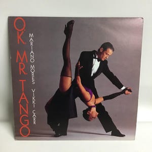 May include: A black and white photo of a couple dancing tango. The woman is wearing a purple top and a black skirt. The man is wearing a black tuxedo. The text "OK. MR. TANGO MARIANO MORES VIKKI CARR" is printed on the album cover.