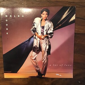 May include: A vinyl record album cover featuring Melba Moore. The cover is a photo of Melba Moore wearing a white and black patterned jacket and white pants. The text "Melba Moore" is in large letters at the top left of the cover. The text "a lot of love" is at the bottom right of the cover.