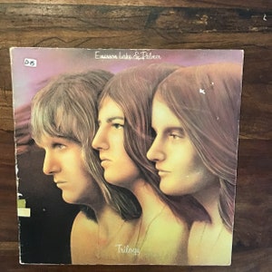 May include: A vintage vinyl record album cover featuring a photo of three men with long hair. The album is titled "Trilogy" by Emerson, Lake & Palmer.