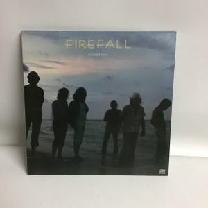 May include: A black and white album cover for the band Firefall. The album cover features a silhouette of a group of people standing on a beach at sunset. The album title "Firefall" and the album name "Undertow" are printed in yellow on the cover.