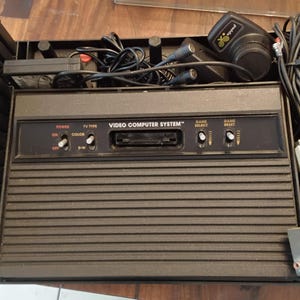 Puede incluir: Una consola Atari Video Computer System negra con un panel frontal de ranuras horizontales, una ranura central para cartuchos e interruptores de palanca para encendido, tipo de TV, selección de juego y reinicio de juego.