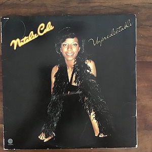 May include: A black and white vinyl record cover featuring a woman wearing a black dress and a feather boa. The album title is "Natalie Cole" and "Unpredictable" in gold lettering.