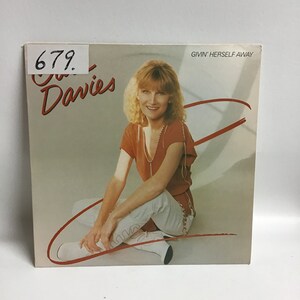 May include: A vinyl record album cover featuring a woman with blonde hair sitting on the floor wearing a red shirt and white jeans. The album title is "Givin' Herself Away" and the artist is "Gail Davies".