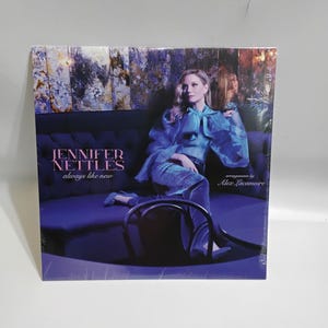 May include: A square album cover featuring Jennifer Nettles. The album title is "always like new." Jennifer is wearing a blue outfit and sitting on a blue couch. The album cover has a purple and blue color scheme.