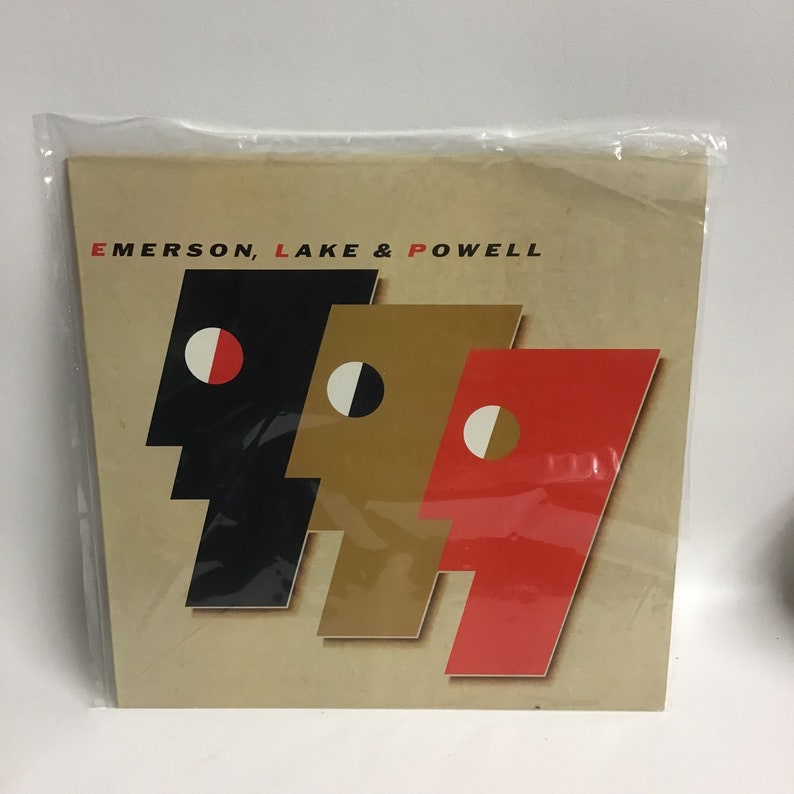 May include: A vintage record album cover featuring the band Emerson, Lake & Powell. The cover art is a stylized design of three faces in black, gold, and red.
