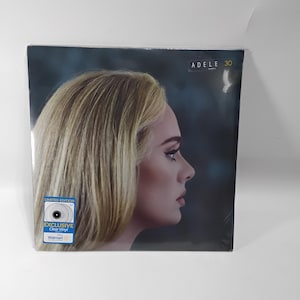 May include: A vinyl record of Adele's album "30" in a clear plastic sleeve. The album cover features a profile portrait of Adele with blonde hair. A sticker on the front reads "Limited Edition Exclusive Clear Vinyl" and includes the Walmart logo.