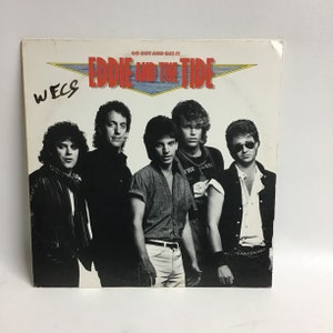 May include: A black and white photo of a band called "Eddie and the Tide" on a white album cover. The album cover has the text "Go Out and Get It" and the band's name in red and blue. The band members are all wearing casual clothing and are looking at the camera.