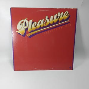 May include: A vintage vinyl record album cover with a red background. The word "Pleasure" is in large, stylized gold lettering with purple accents. The text "SPECIAL THINGS" is below in smaller gold letters.