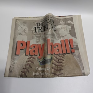 May include: A newspaper front page from The Tampa Tribune, dated March 31, 1998, with the headline "Play ball!" in large orange letters. The image features baseball players and a baseball graphic, with text that reads "It's Opening Day for the Devil Rays."