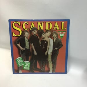 May include: A red and blue record album cover with the band "SCANDAL" in large yellow letters. The band members are pictured in black and white, standing in front of a red background. The album title is "Love's Got A Line On You".