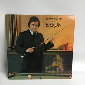 May include: A vinyl record cover featuring Johnny Cash, holding a pool cue, with the album title "The Baron" in large, bold letters.
